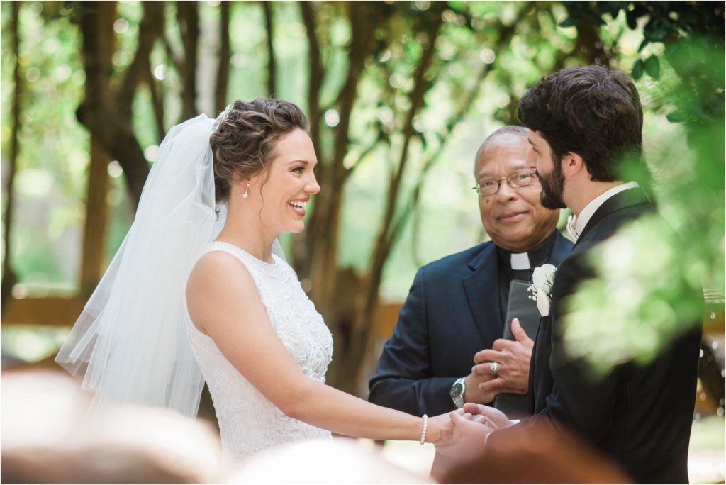 Wedding ceremony taking place outdoors with officiant and couple exchanging vows under green trees.