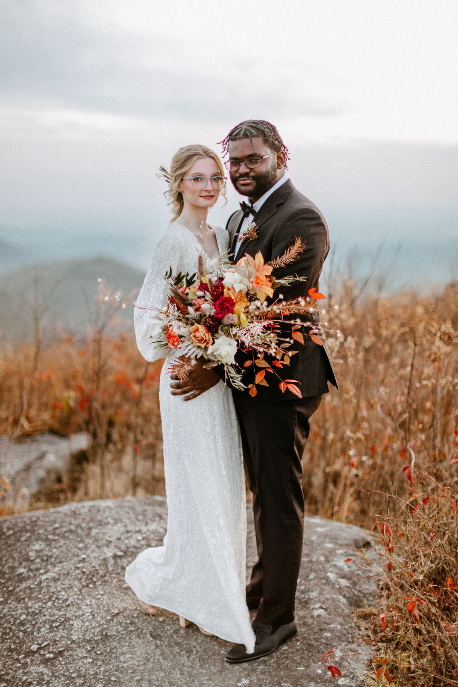 A couple poses with a vibrant autumn bouquet on a mountaintop during their wedding.