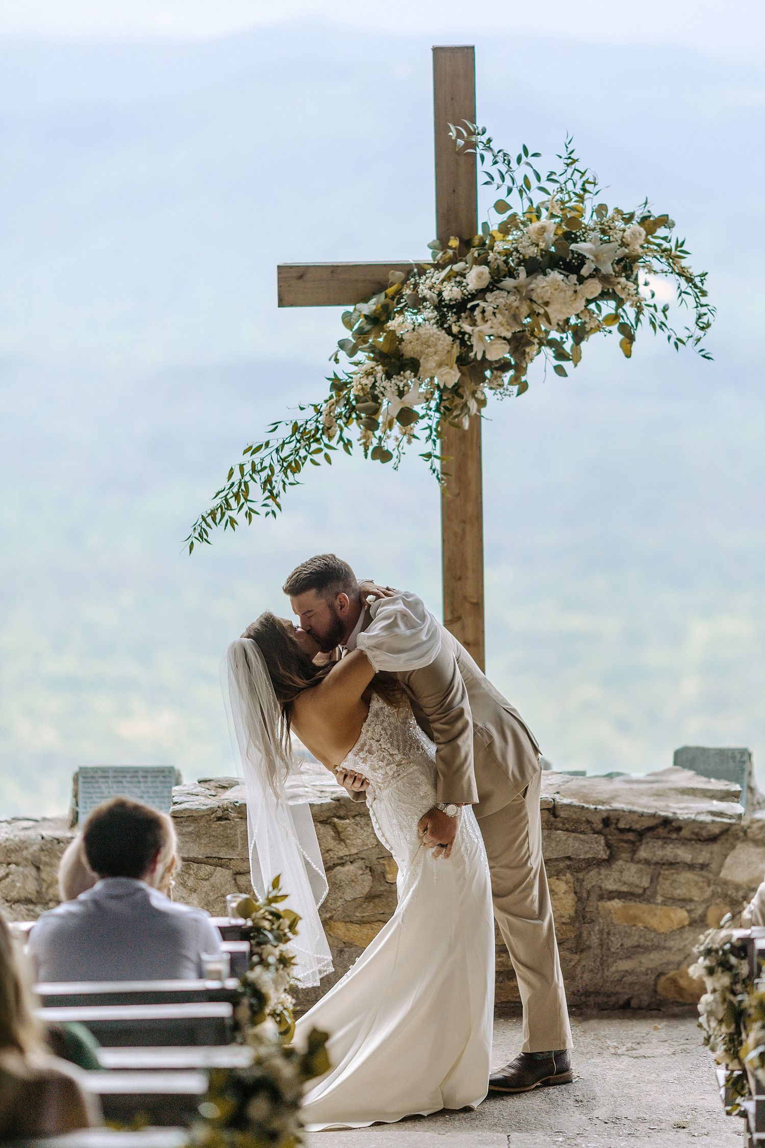 Romantic wedding moment under decorated wooden cross with mountain vista backdrop.