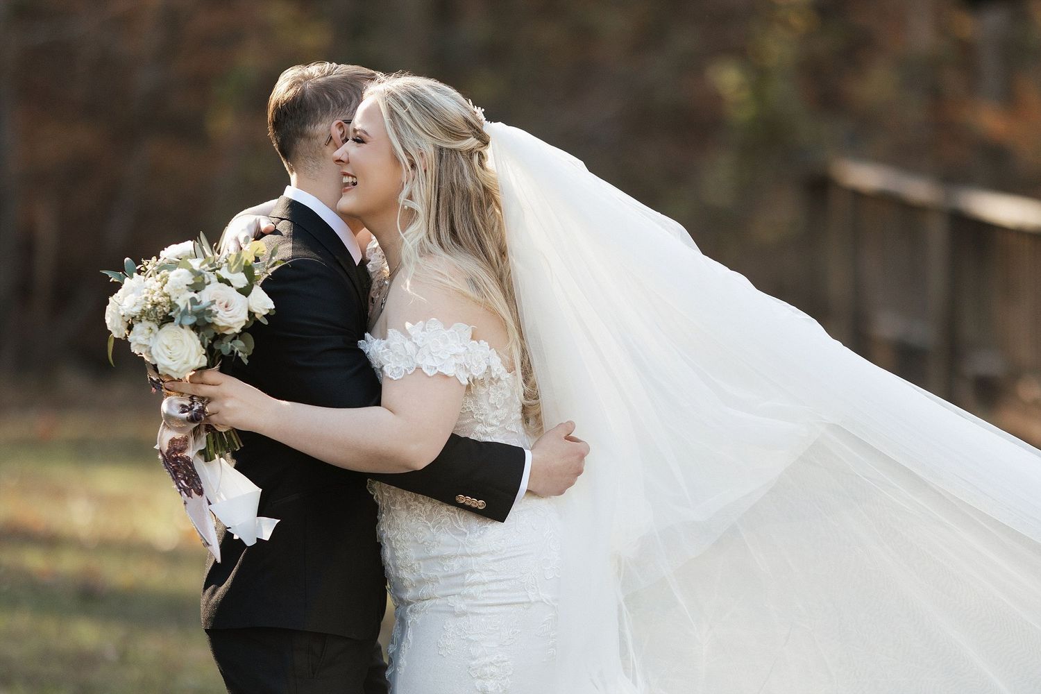 A romantic outdoor wedding moment showing a couple embracing while holding a white floral bouquet.