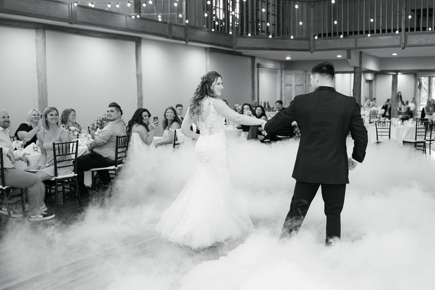 A couple shares their first dance on a misty dance floor at a wedding reception as guests look on from their tables.
