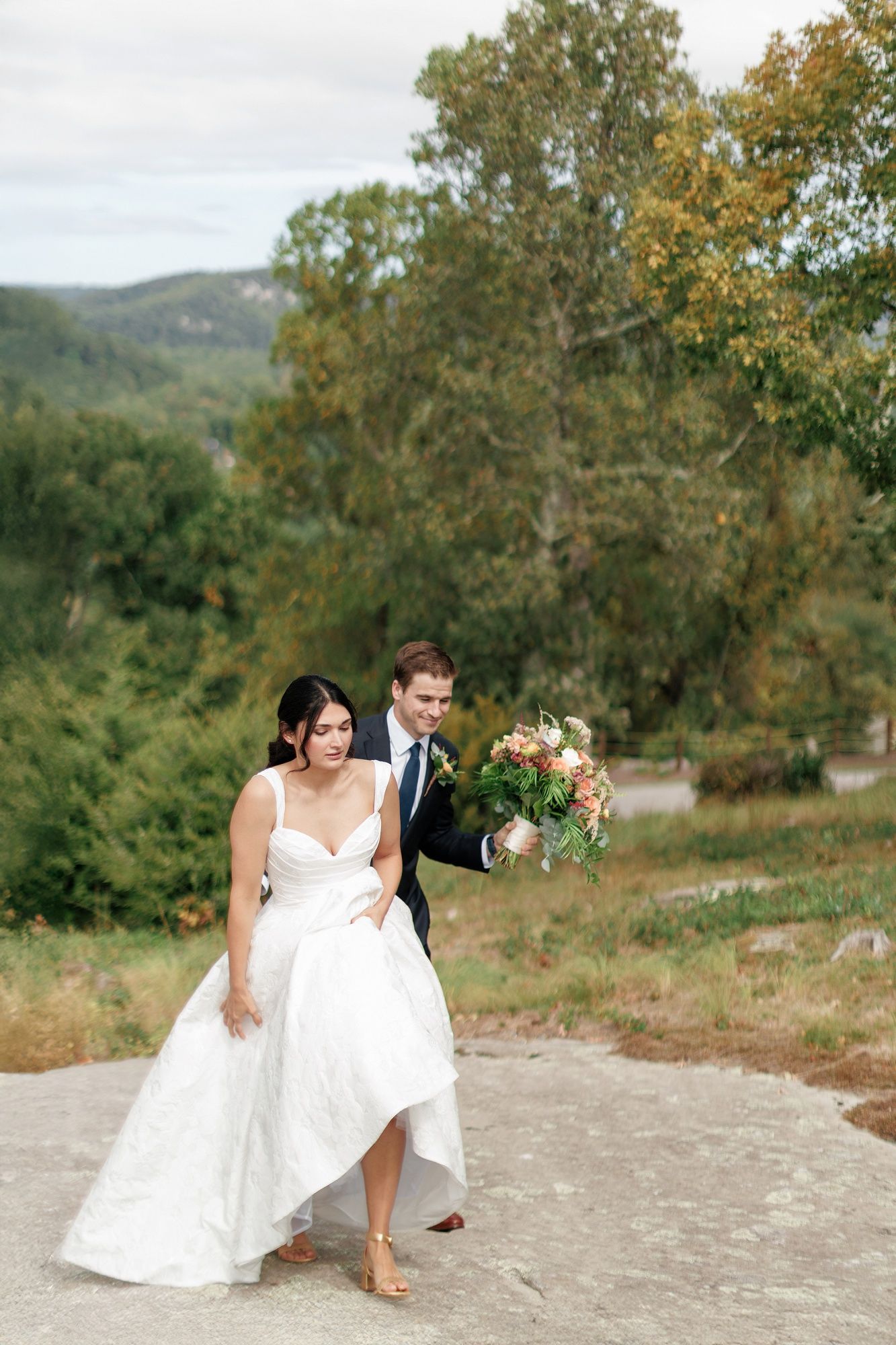 Bride and groom playfully pose together on a stone surface with autumn trees and mountains in the background.