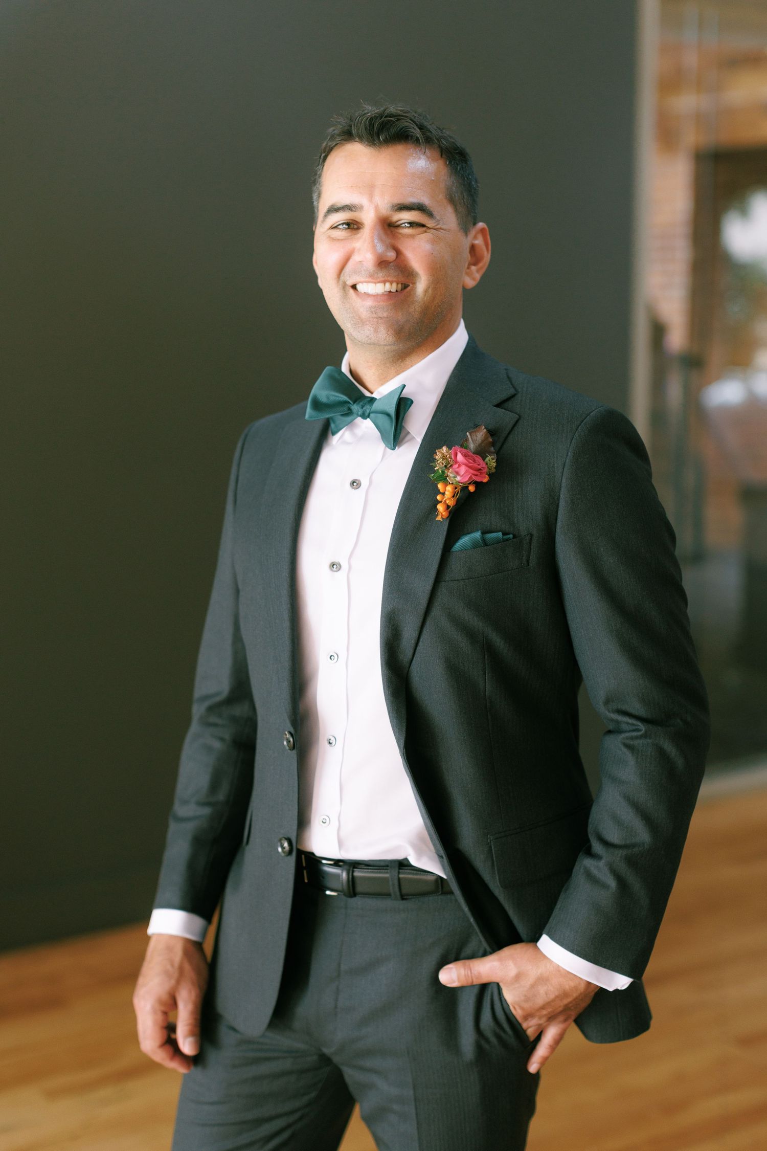 Smiling groom in dark suit with green bow tie and floral boutonniere poses confidently at wedding venue.