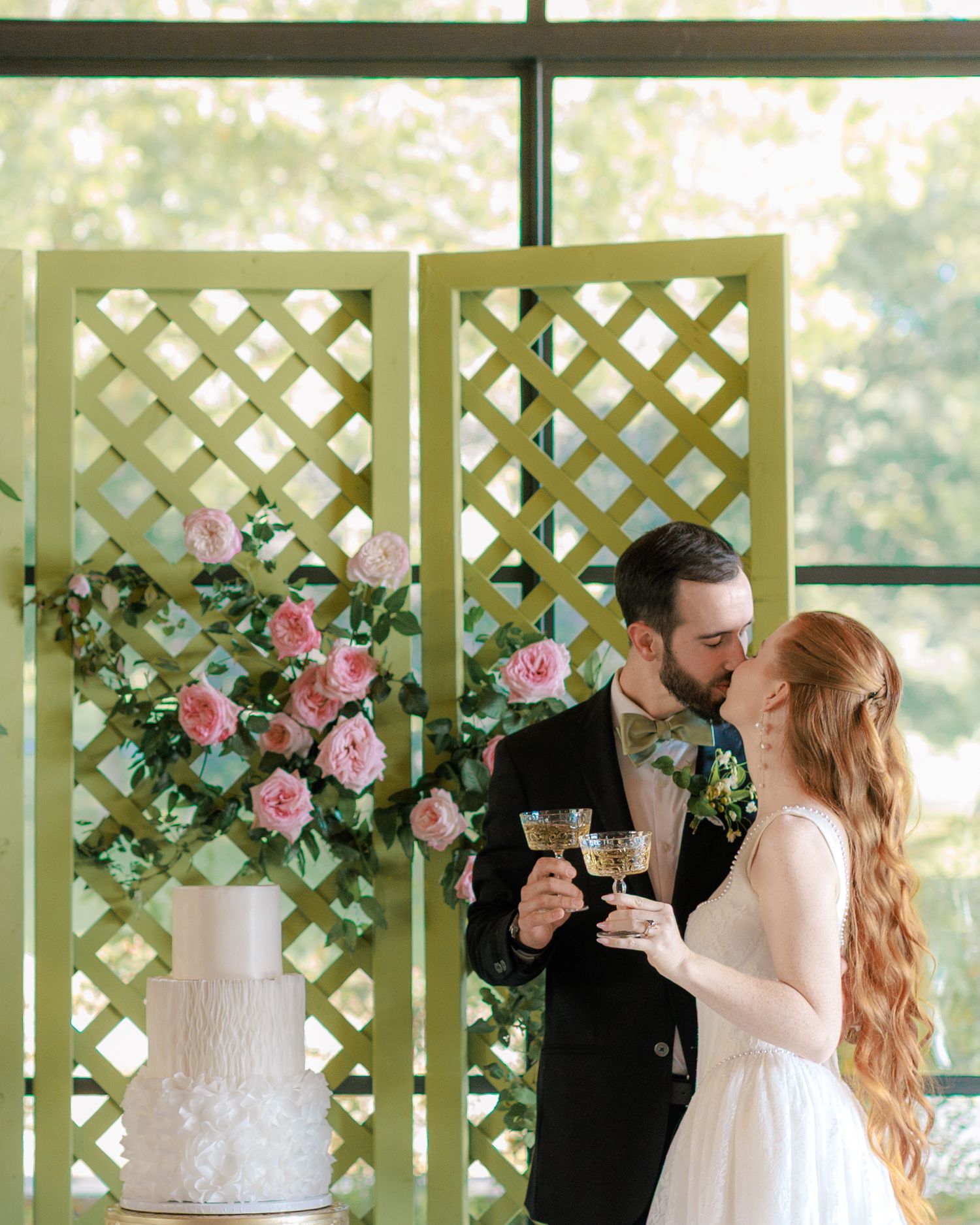 Bride and groom share a kiss while toasting with champagne glasses in front of a green lattice backdrop with pink roses.