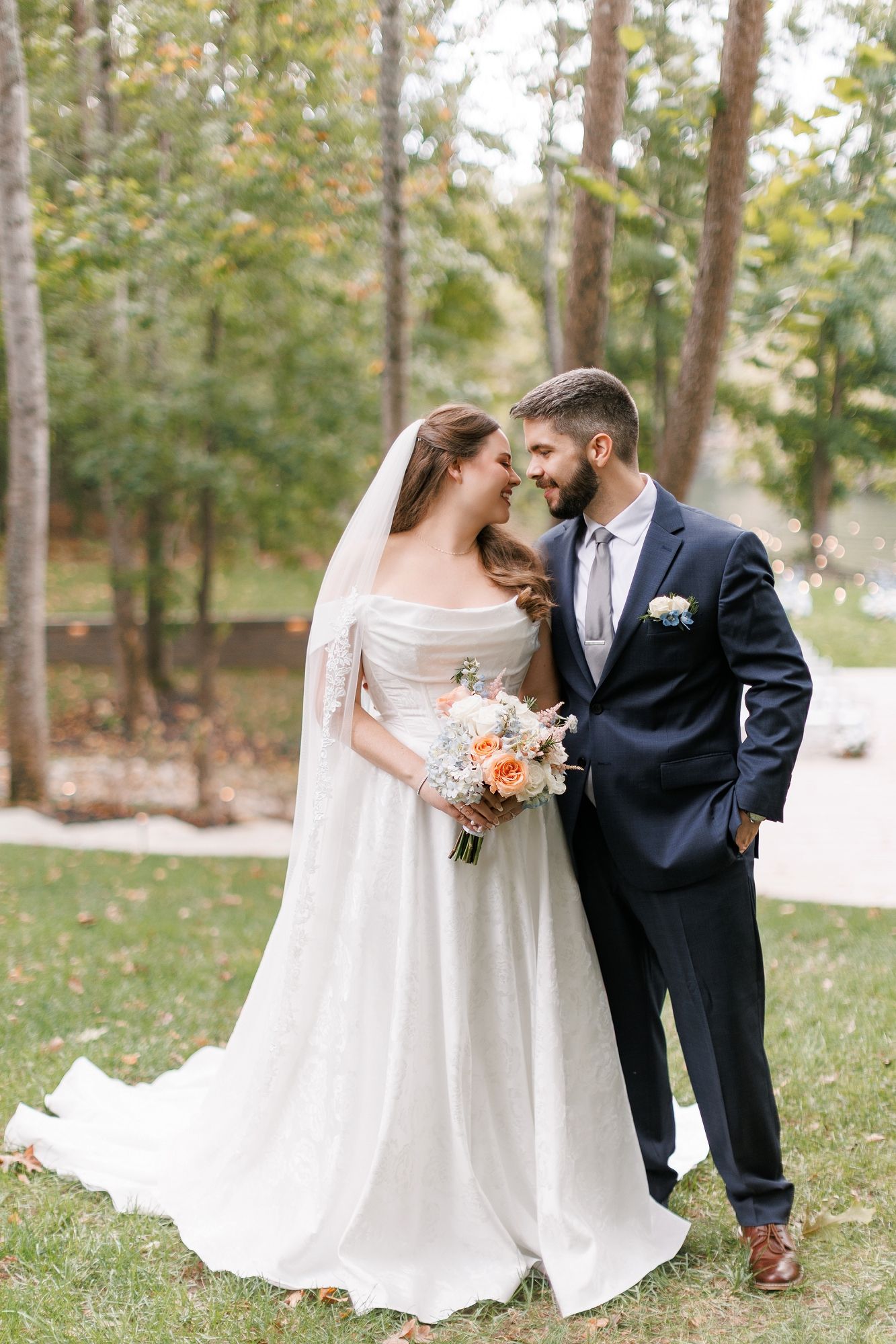 Bride and groom share a romantic moment outdoors, she in a white gown with veil, he in a navy suit.