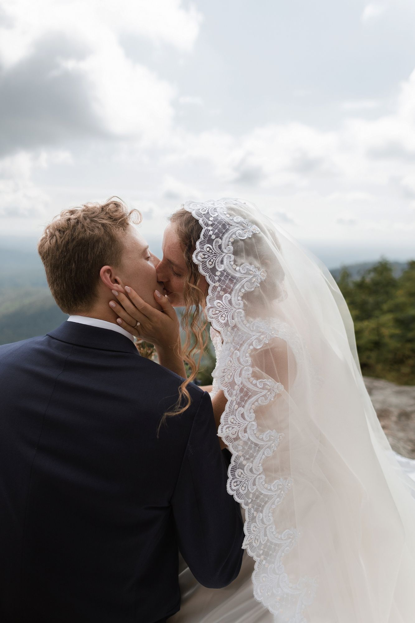 Bride and groom share a romantic kiss on a mountain overlook, her lace veil flowing in the breeze.