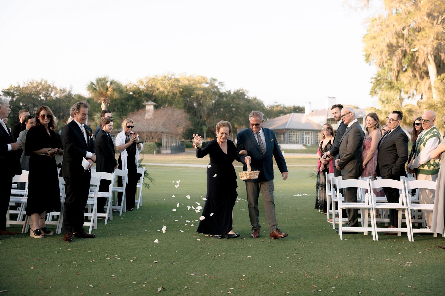Wedding guests seated in white chairs on a lawn for an outdoor ceremony at sunset.