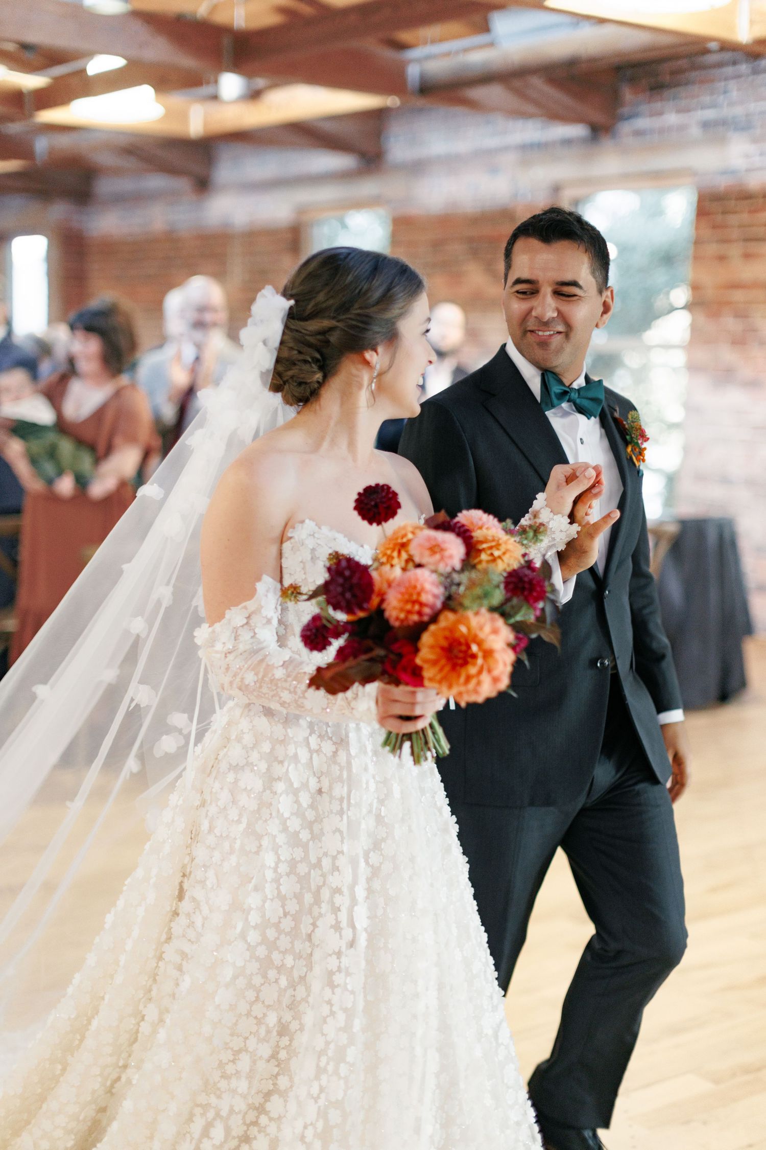Bride in lace gown with colorful dahlia bouquet dances with groom in black tuxedo at elegant wedding reception.