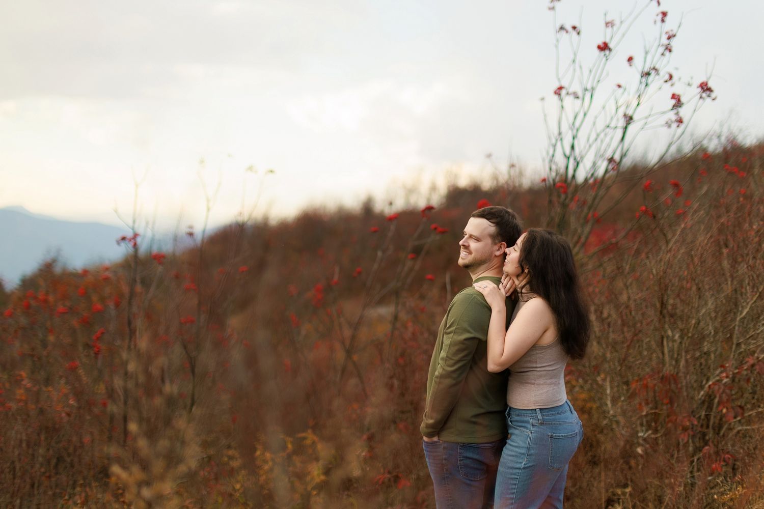 A couple embraces while standing on a mountainside during autumn with red wildflowers in the background.