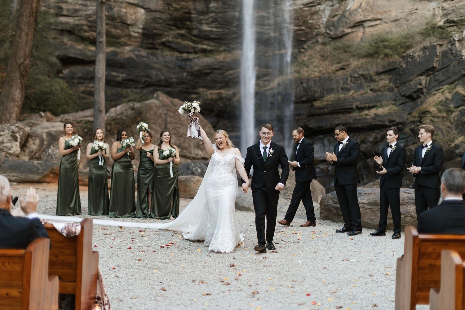 A wedding party stands in formation against a dramatic waterfall backdrop at an outdoor ceremony venue.