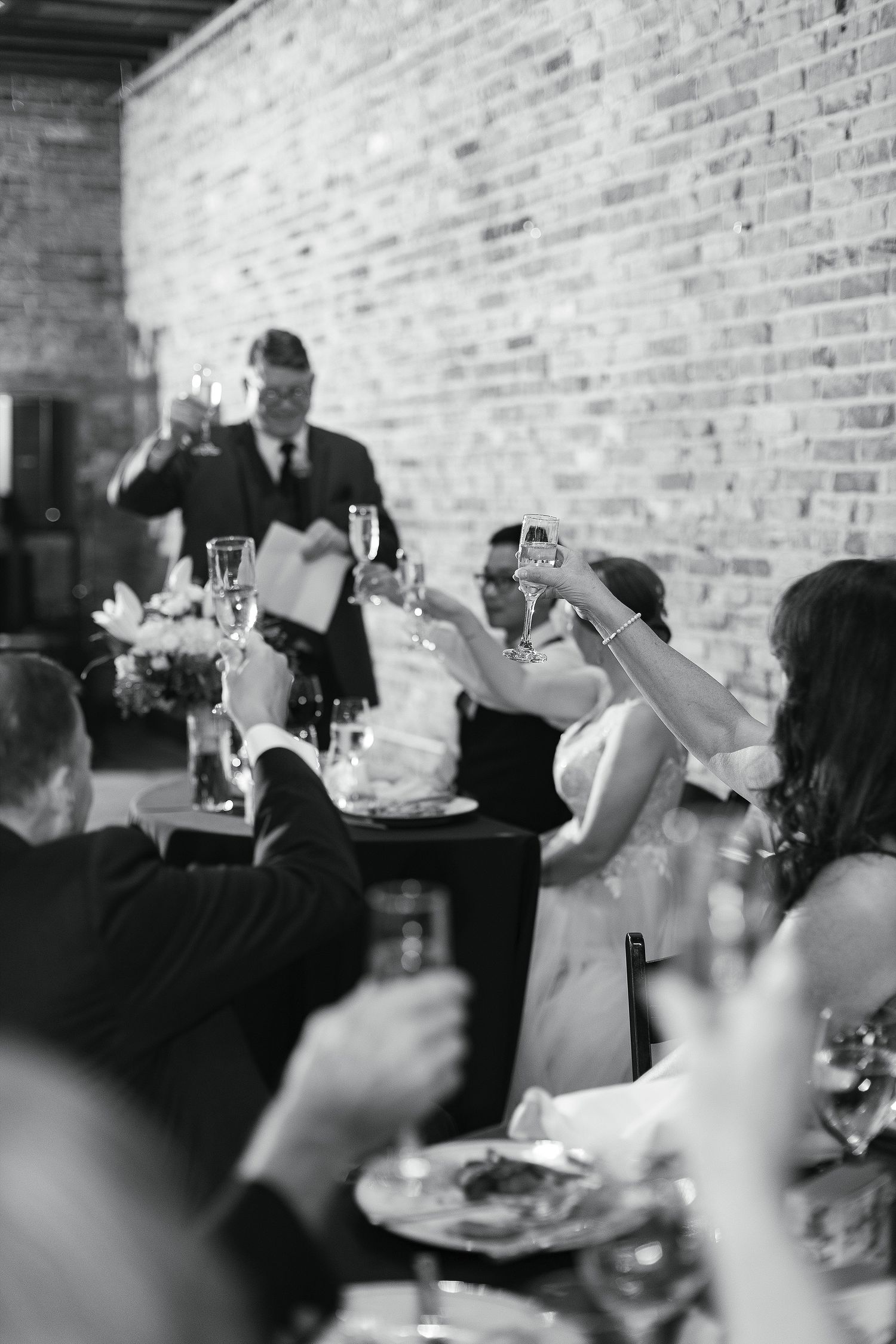 Wedding guests raise glasses in a toast at reception dinner with exposed brick wall backdrop.