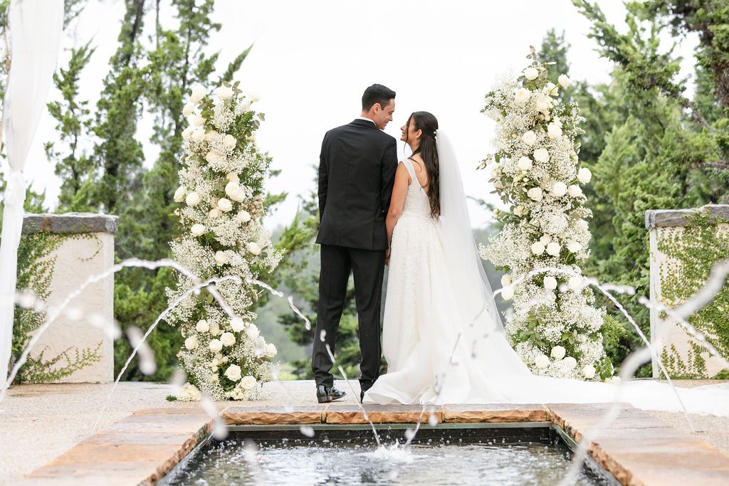 A wedding couple stands at an elegant outdoor ceremony site with white floral arrangements and a reflecting pool.