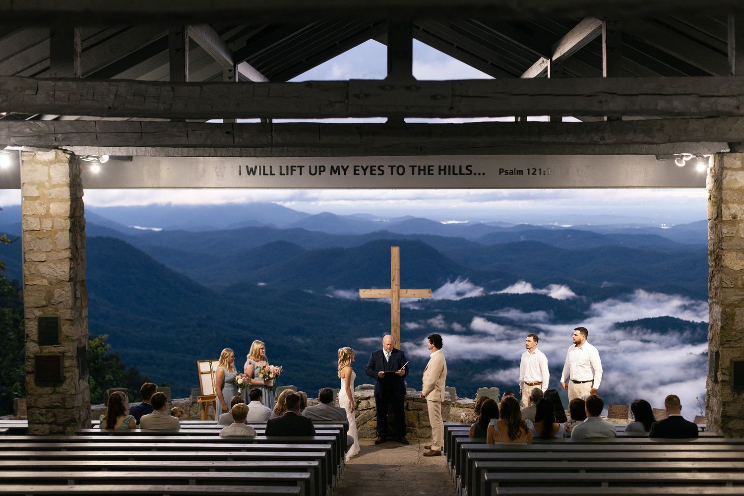 Visitors gather at a mountain overlook platform with a cross, viewing misty blue ridges and valleys at sunset.