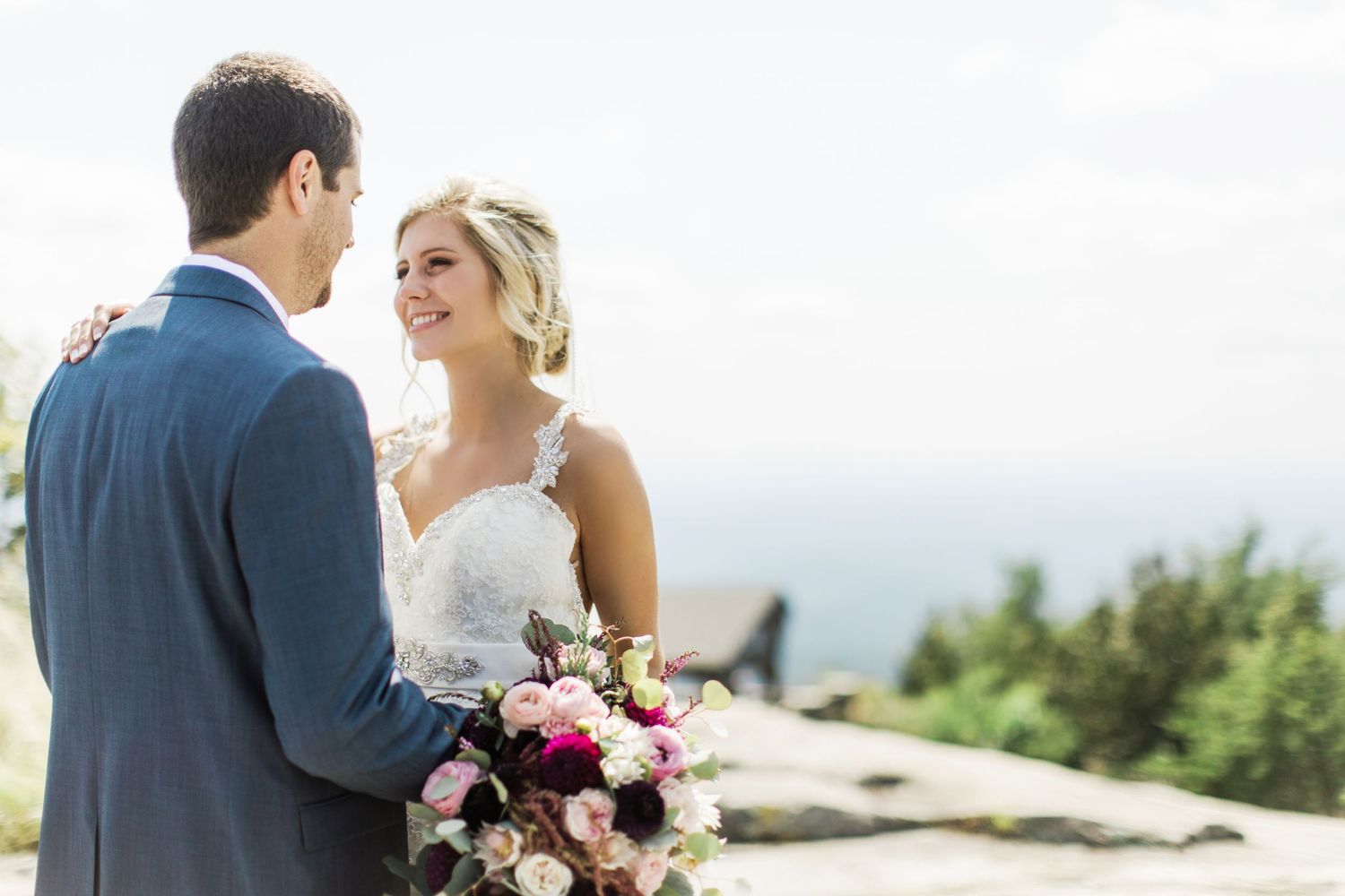 A bride and groom share a moment on a scenic mountaintop with a colorful floral bouquet after their Cliffs at Glassy Chapel Wedding.