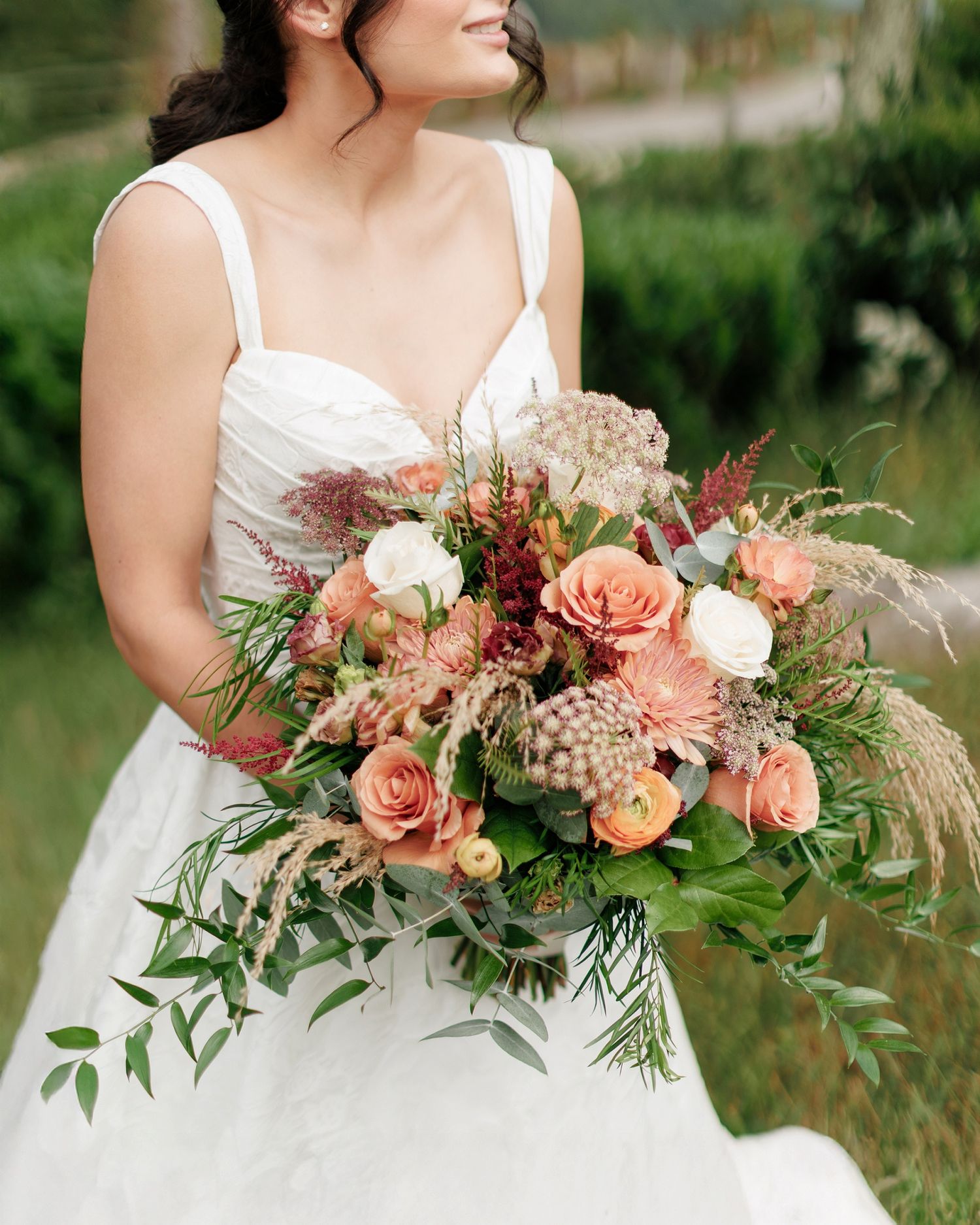 A wedding bouquet with coral roses, peach flowers, and greenery held against a white dress in an outdoor setting.