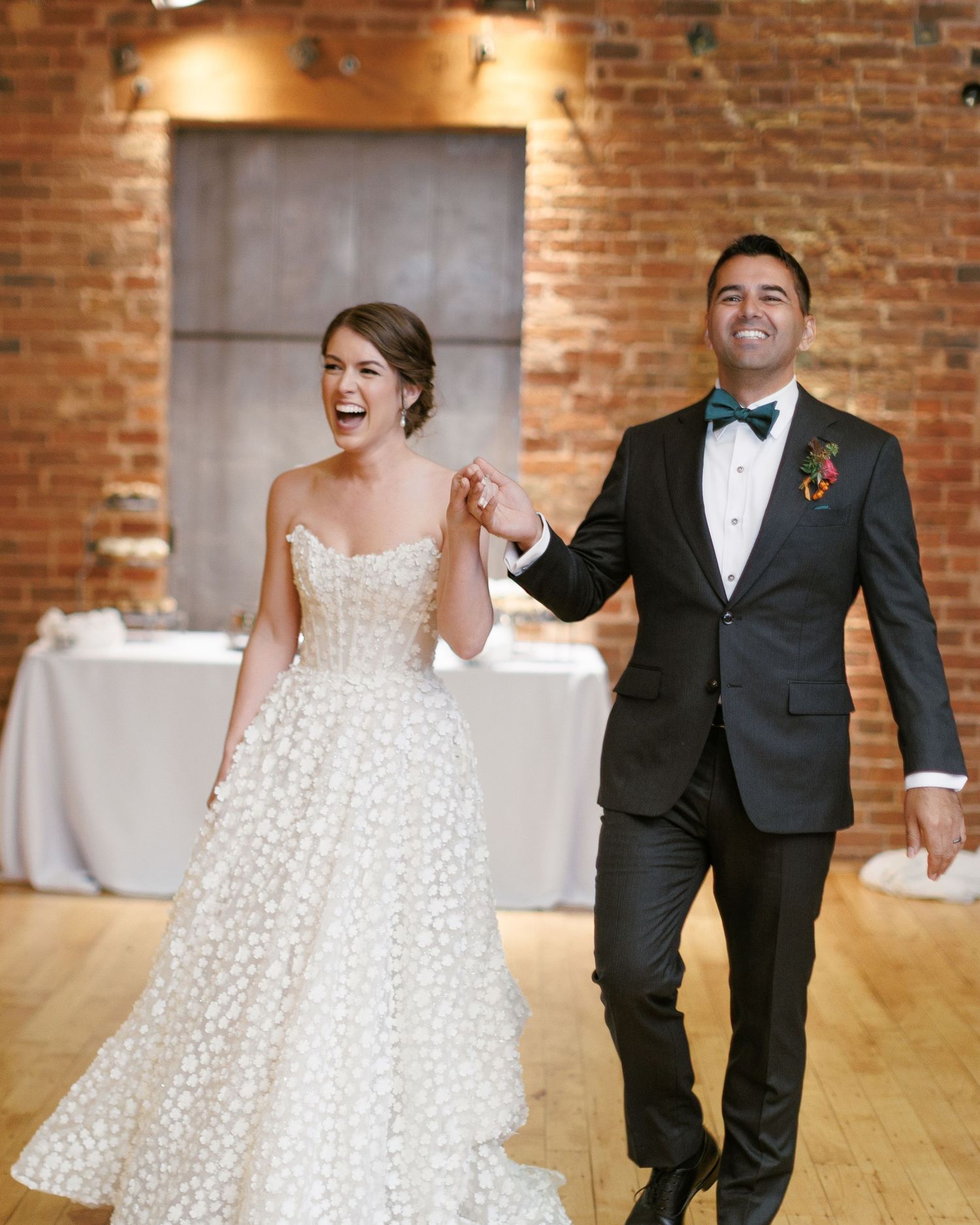 Smiling bride in white floral gown and groom in black tuxedo with bow tie make grand entrance at wedding reception.