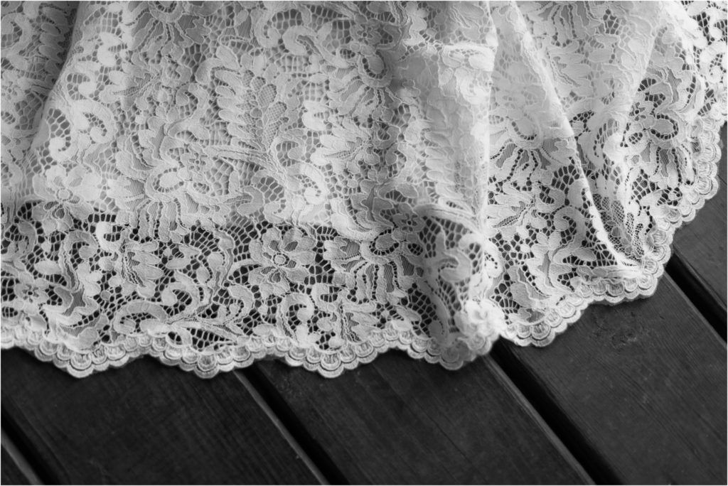 Close-up detail of intricate lace pattern on wedding dress hem against wooden deck.
