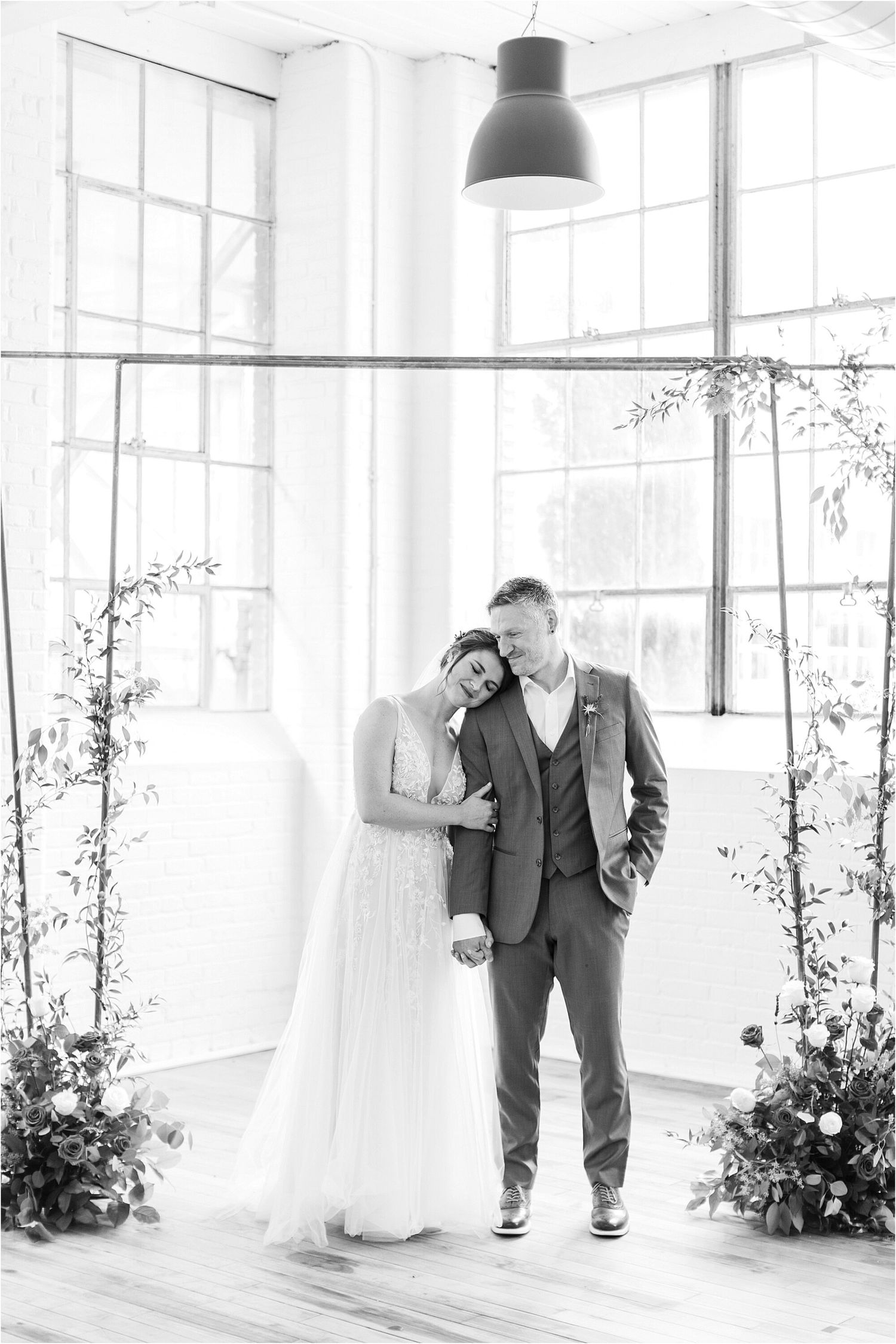 Black and white photo of couple in wedding attire standing by bright windows with floral arrangements.