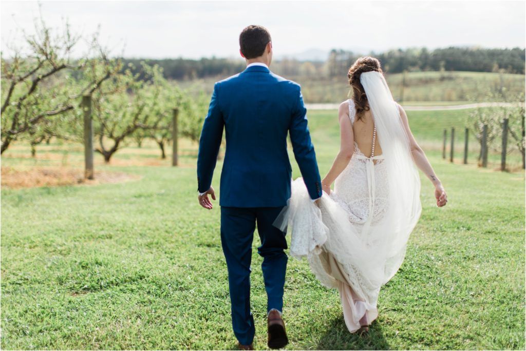 A couple on their wedding day walk through a scenic orchard with rolling hills in the background.