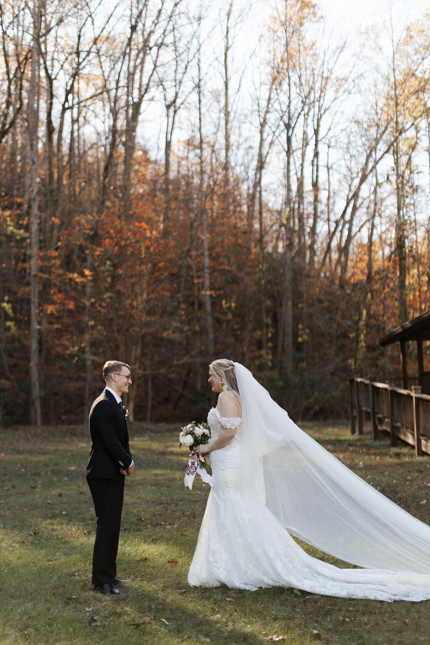 A couple shares their wedding day moment outdoors in a wooded autumn setting with a flowing white cathedral veil.