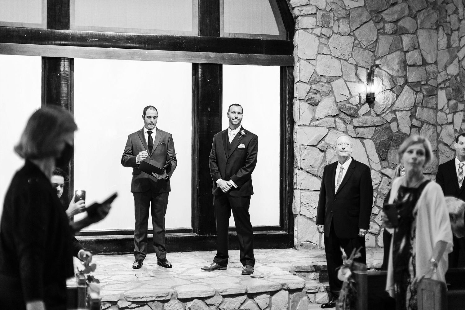 Wedding ceremony attendees gather in a stone chapel with large windows and natural lighting.