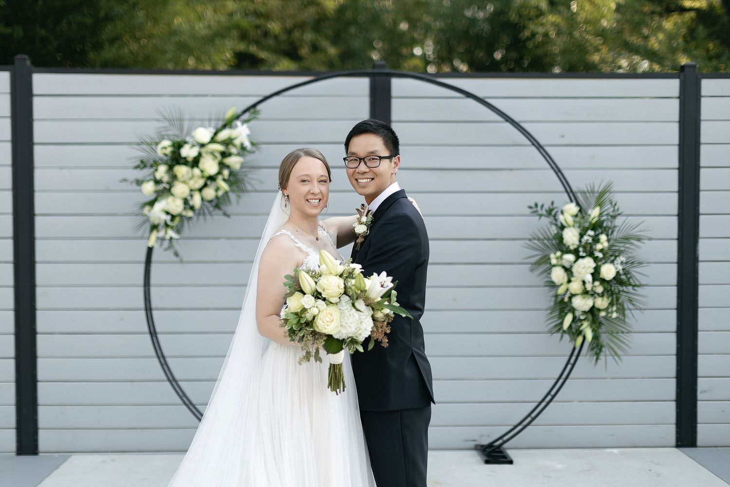 Couple poses in front of modern circular wedding arch with white floral arrangements.