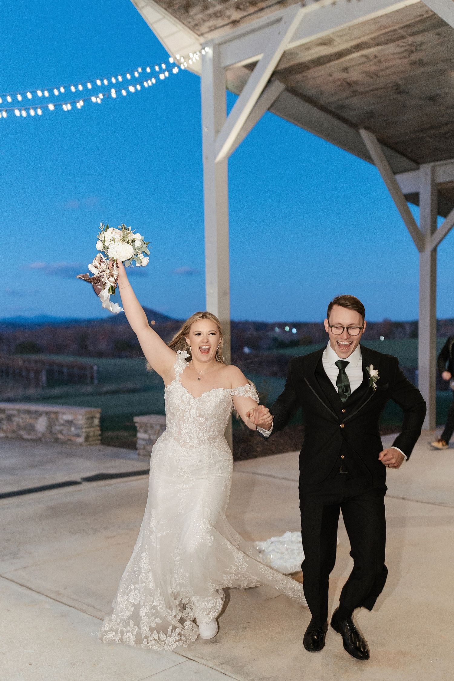 A couple celebrates their wedding at a scenic outdoor venue with string lights and mountain views in the background.
