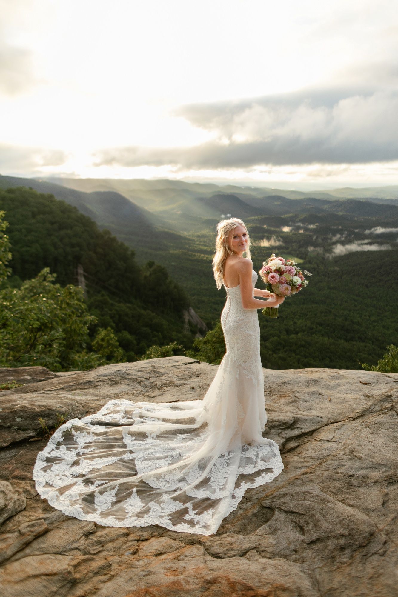 A bride in a flowing lace wedding gown holds a pink bouquet while standing on a scenic mountain overlook at sunset.