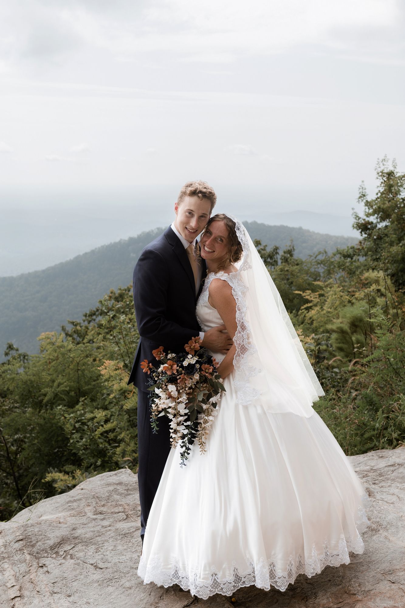 Bride and groom posing on a rocky overlook with mountain views, bride holding autumn bouquet in white lace gown with veil.