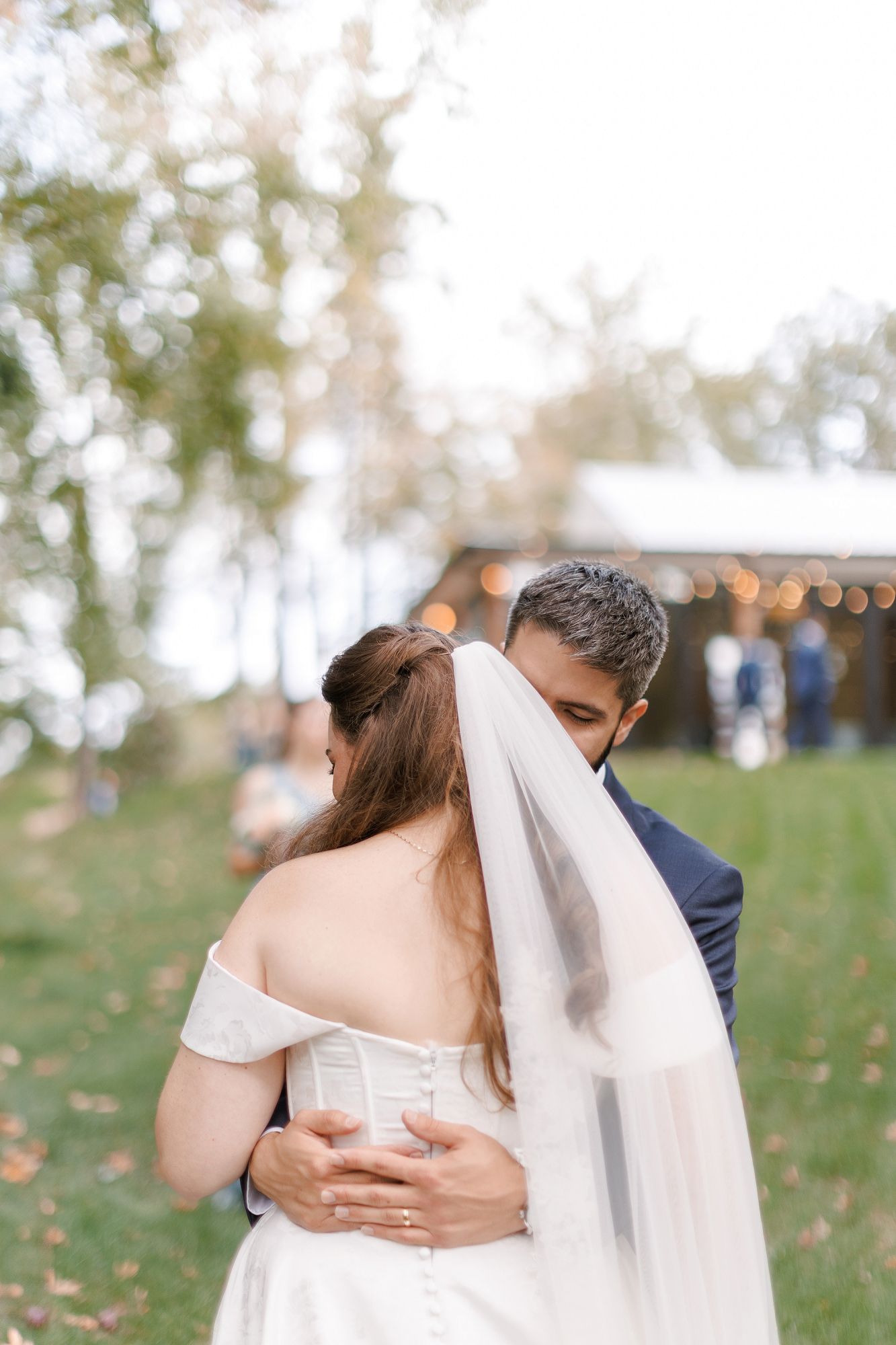 Bride and groom share tender embrace outdoors, groom nuzzling bride's veil, with soft bokeh lights in background.