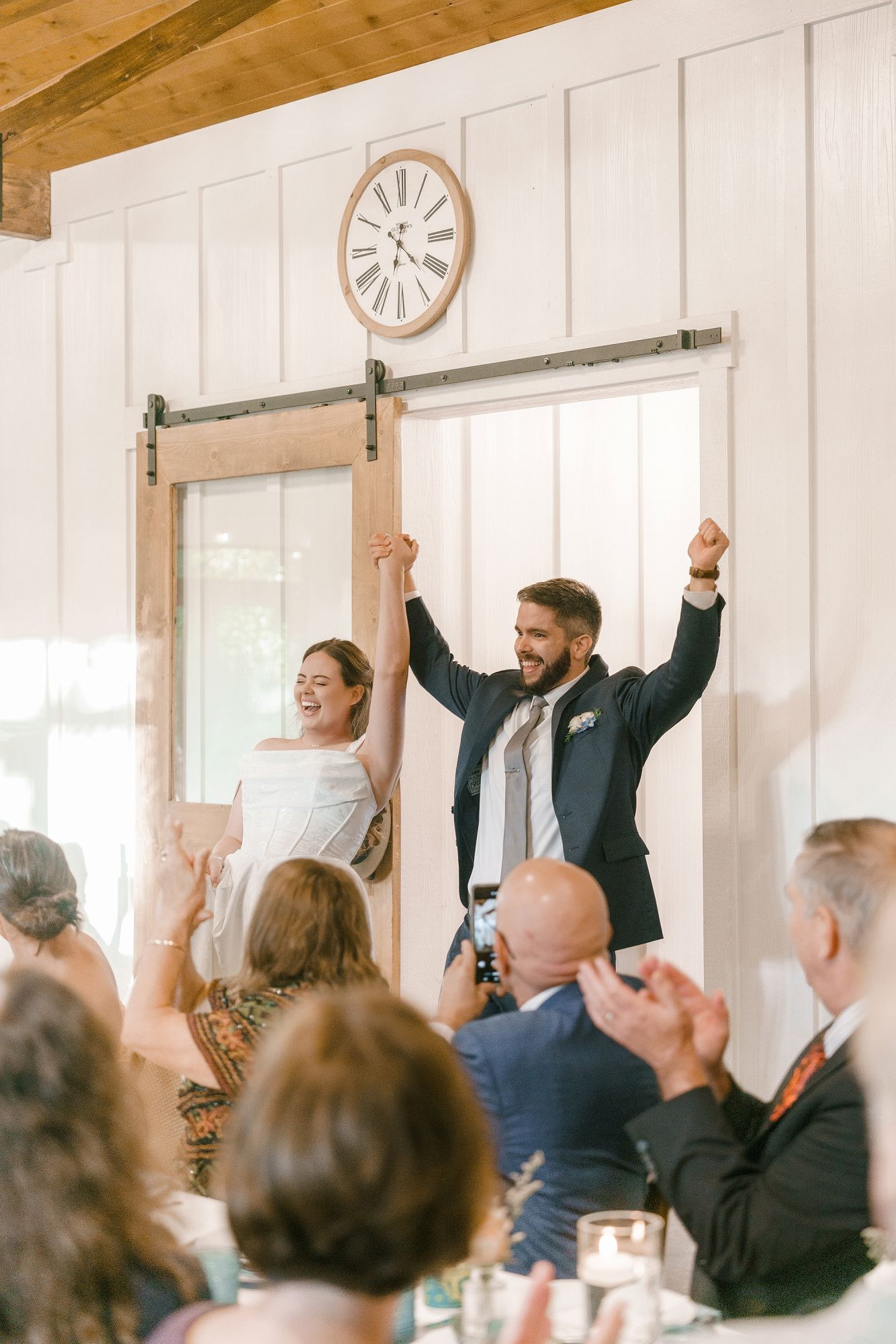 Bride and groom raise their hands in celebration as they enter their wedding reception through a rustic barn door.