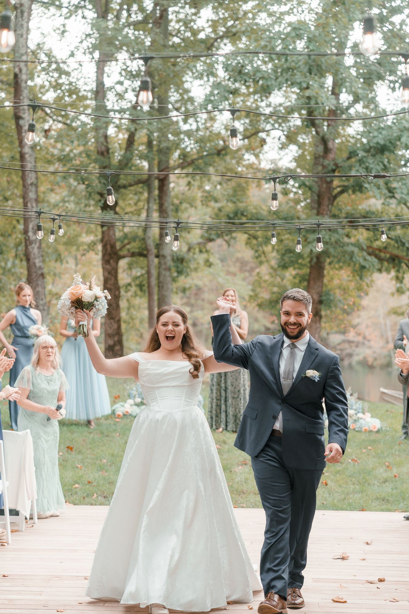 Joyful newlyweds raise hands celebrating as they walk back down the aisle after outdoor woodland wedding ceremony.
