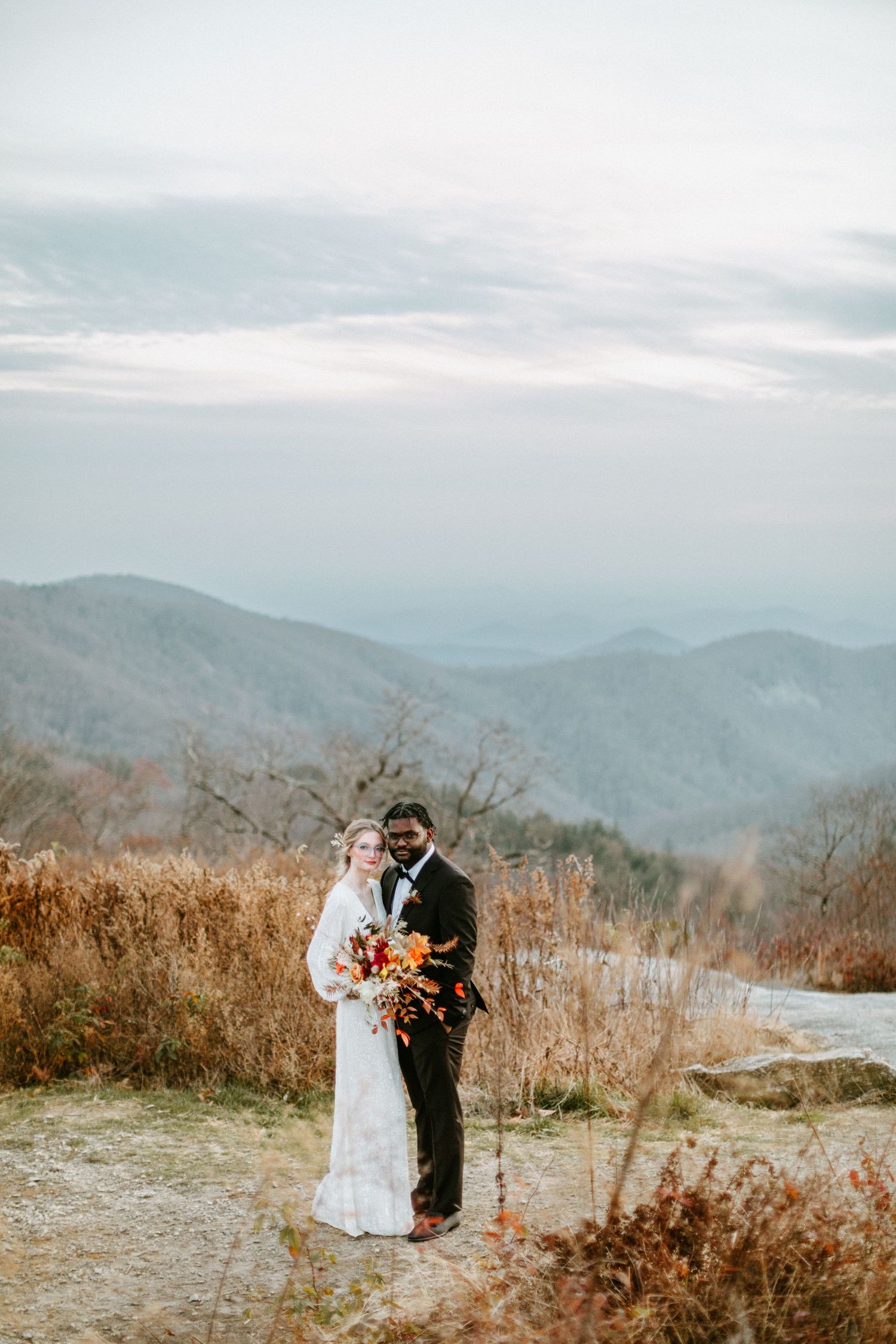 Newlyweds pose together on a mountaintop trail holding orange and red autumn bouquet with mountain vistas behind.