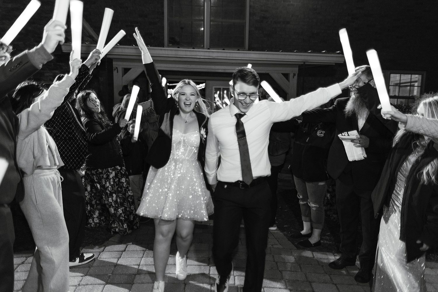 Wedding celebration with couple exiting through a tunnel of guests holding light-up foam sticks at night.