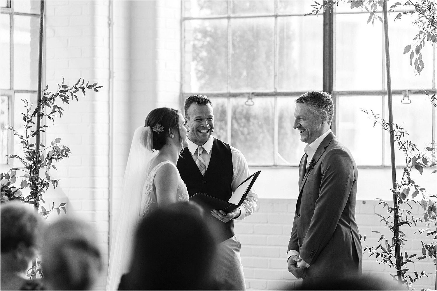A wedding ceremony takes place in a bright sunlit room with tall windows and greenery decorations.