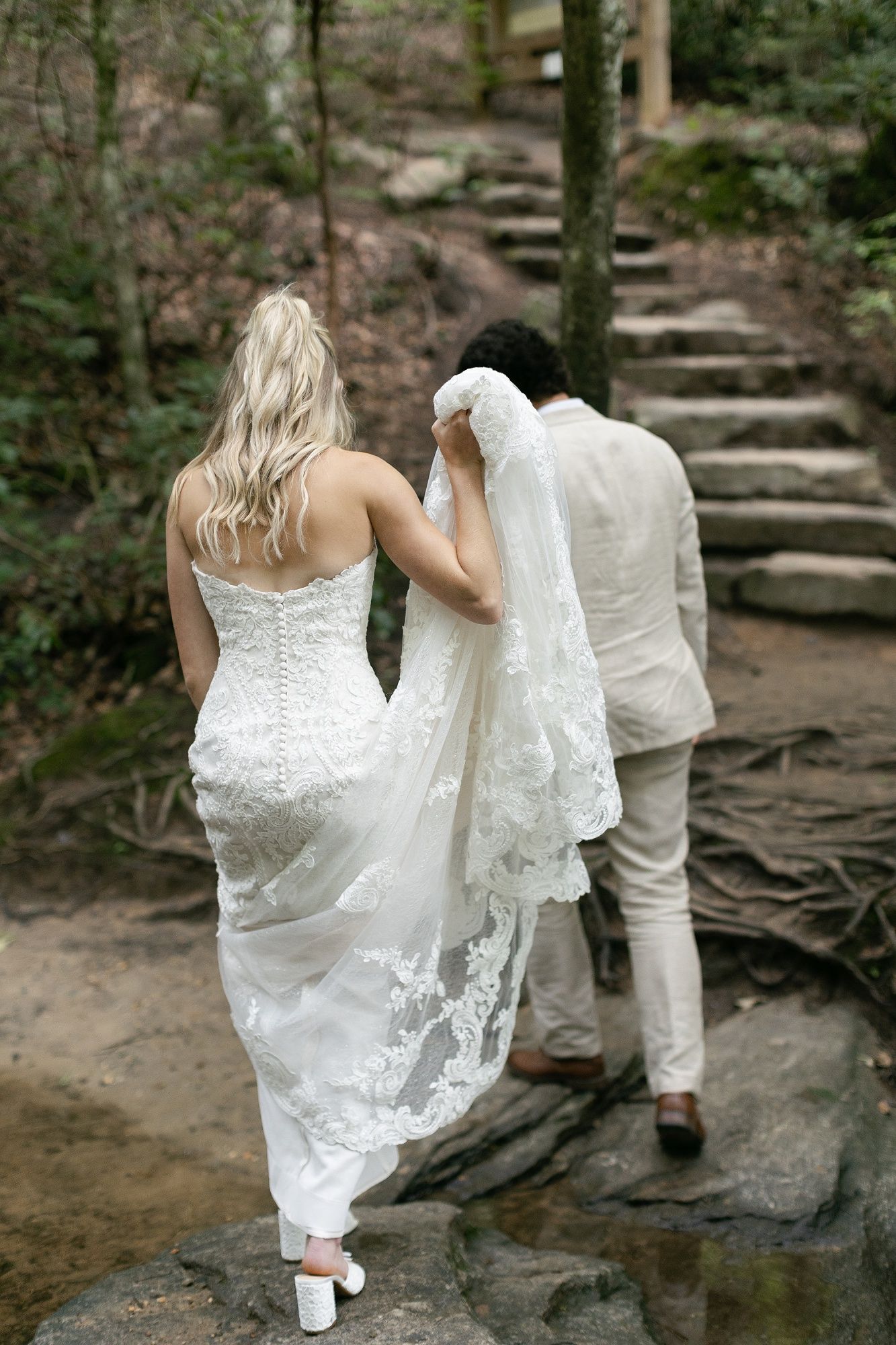 A couple in wedding attire carefully ascending stone steps on a forest trail.