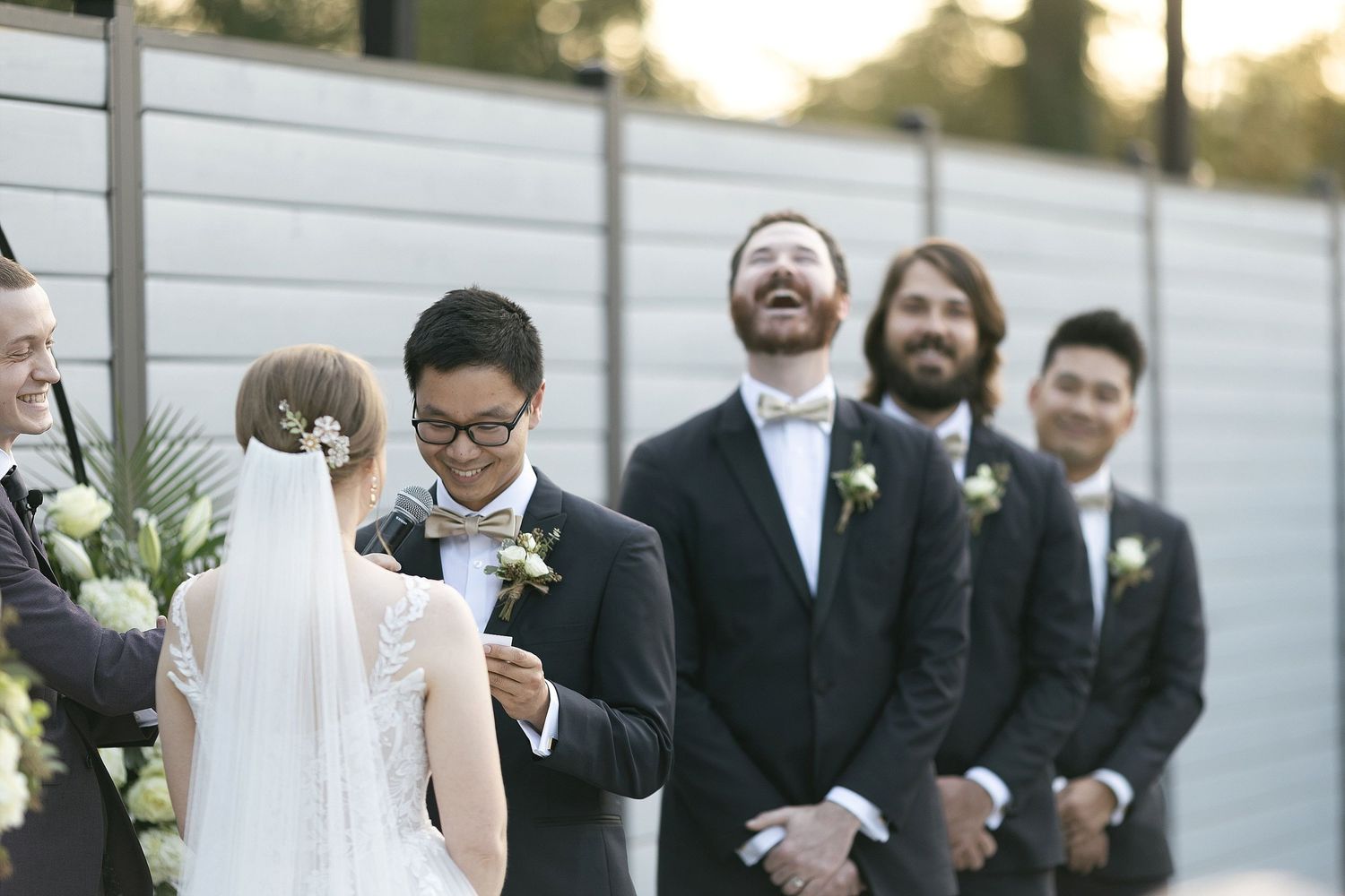 Wedding party members share a joyful moment during an outdoor ceremony with flowers and warm sunset lighting.