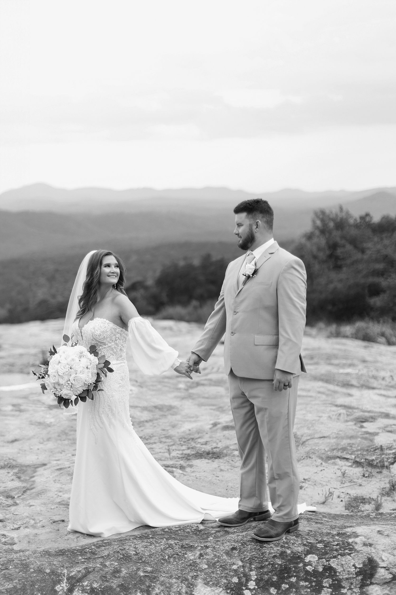Elegant wedding couple poses on rocky mountain outcrop with scenic valley views.