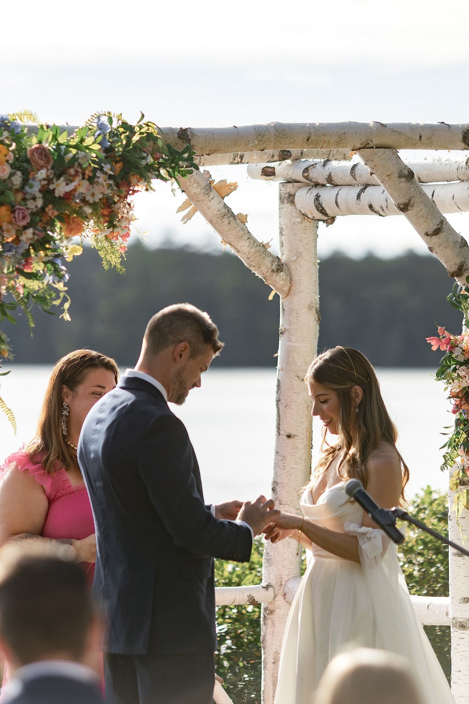 A couple exchanges rings under a birch wedding arch decorated with flowers by a lakeside.