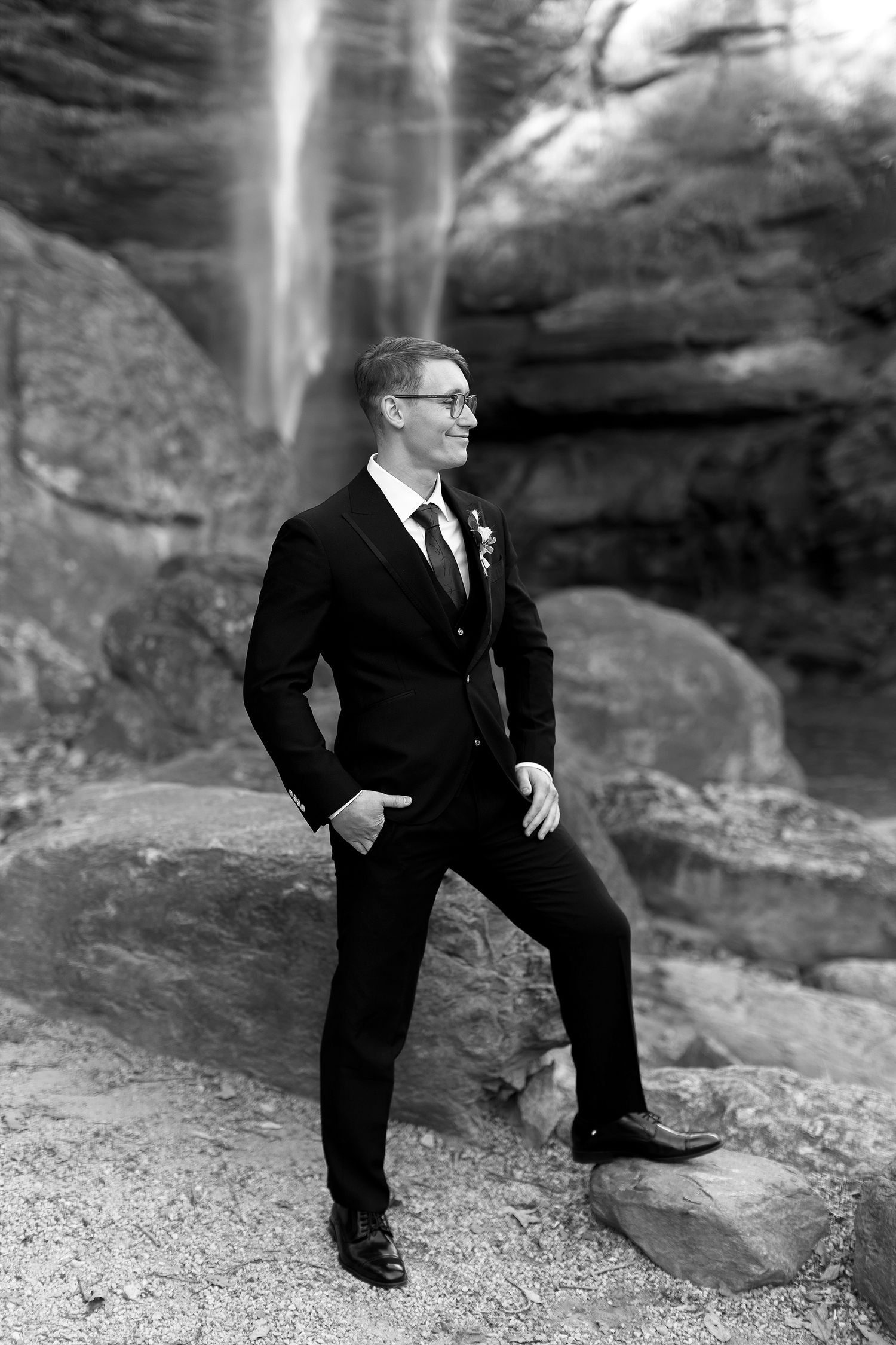 A person in a formal black suit and tie poses confidently on rocky terrain with a waterfall in the background.
