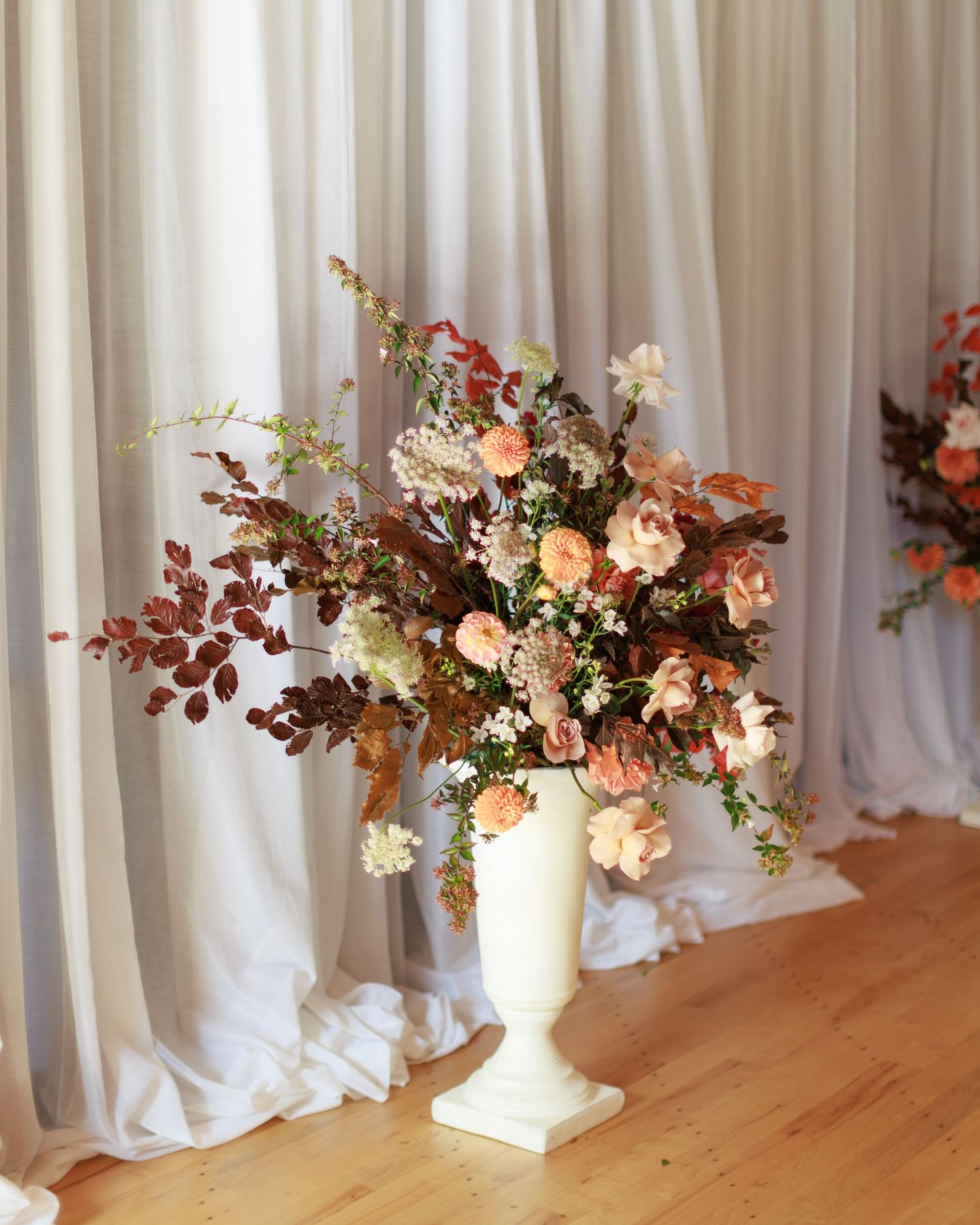 Elegant white urn vase with lush autumn floral arrangement featuring peach roses and burgundy foliage against draped curtains.
