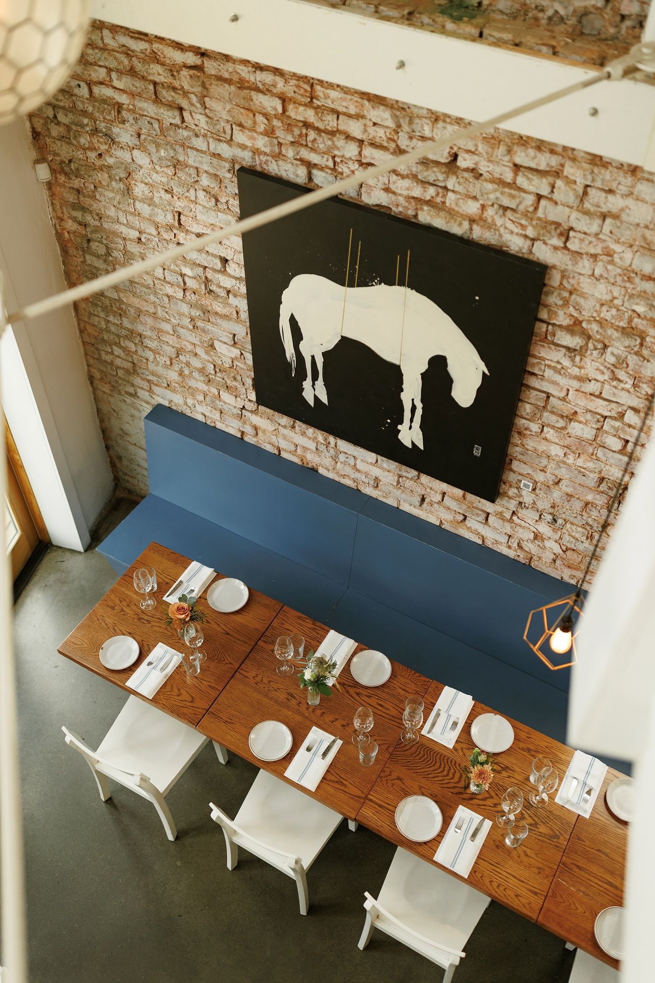 Modern restaurant interior with wooden tables, white chairs and black horse silhouette artwork on exposed brick walls.