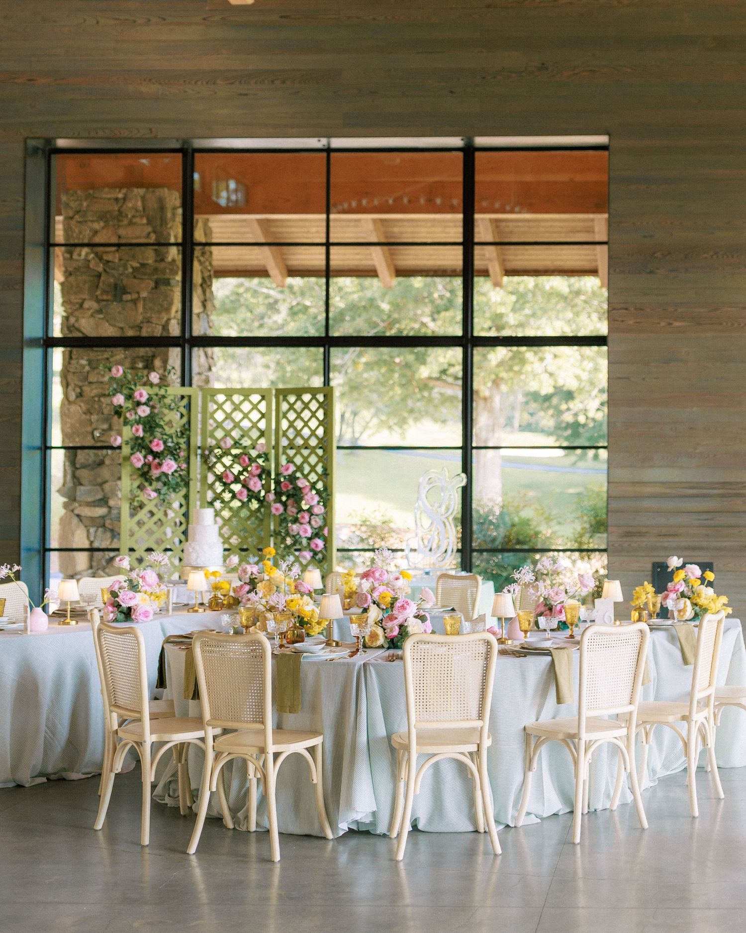 Elegant wedding reception with floral centerpieces, rattan chairs, and large glass windows in a rustic venue.