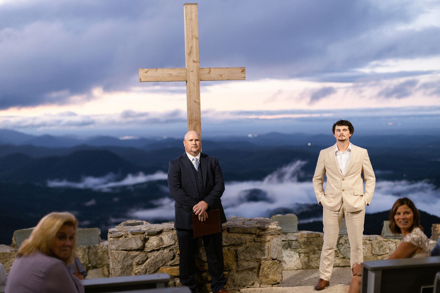 A wooden cross stands atop a mountain overlook with misty blue ridges and dramatic clouds in the background.