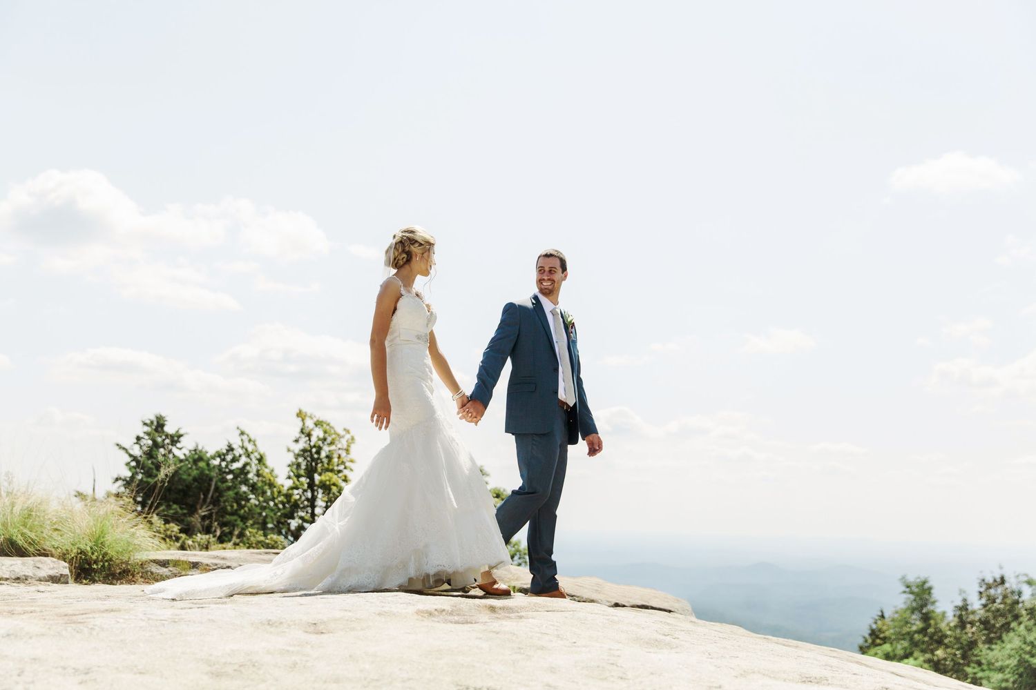 A couple in wedding attire stands together holding hands on top of a mountain overlook on a clear summer day at The Cliffs at Glassy Chapel.
