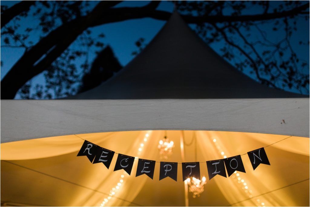 Illuminated wedding tent ceiling with decorative banner and string lights at dusk.