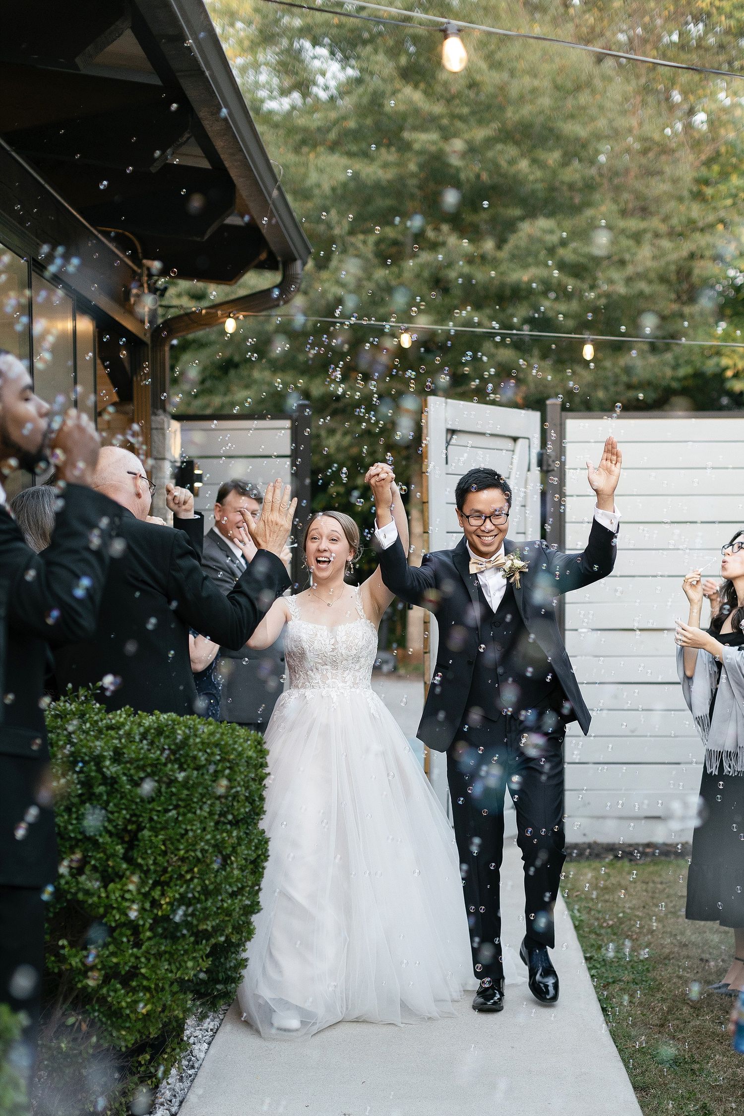 A joyful couple celebrate their wedding ceremony on an outdoor path as guests shower them with bubbles.