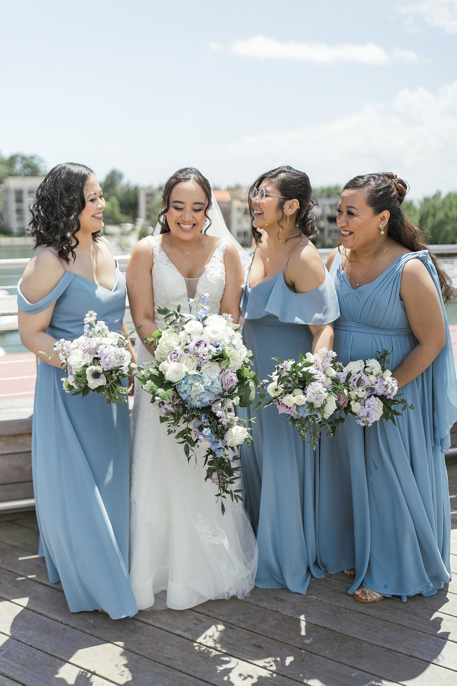 Bride and bridesmaids in light blue dresses share a candid moment on waterfront deck.