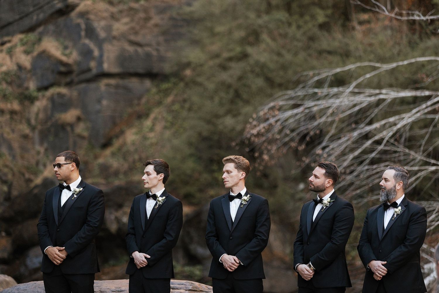 A row of groomsmen in black tuxedos and bowties stand together at an outdoor wedding ceremony.