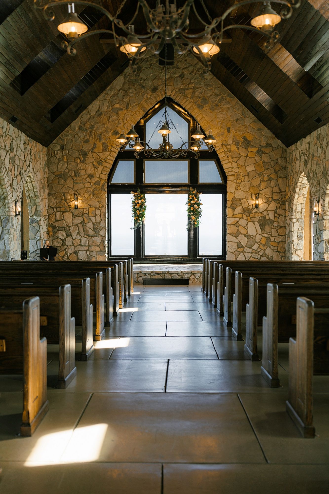 Interior view of a rustic stone chapel with wooden beams and pendant lights creating a dramatic cathedral ceiling.